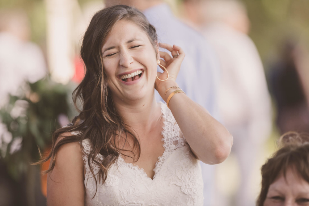 Une mariée heureuse vêtue d'une robe en dentelle rit bruyamment en touchant son oreille, tandis que ses invités sont légèrement flous derrière elle lors d'un événement en plein air.