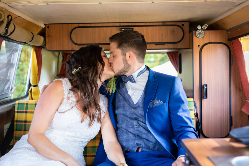 Un couple nouvellement marié s'embrasse dans un camping-car à l'ancienne aux intérieurs confortables et aux décorations à carreaux. La mariée porte une robe en dentelle blanche et le marié porte un costume bleu avec une décoration florale.