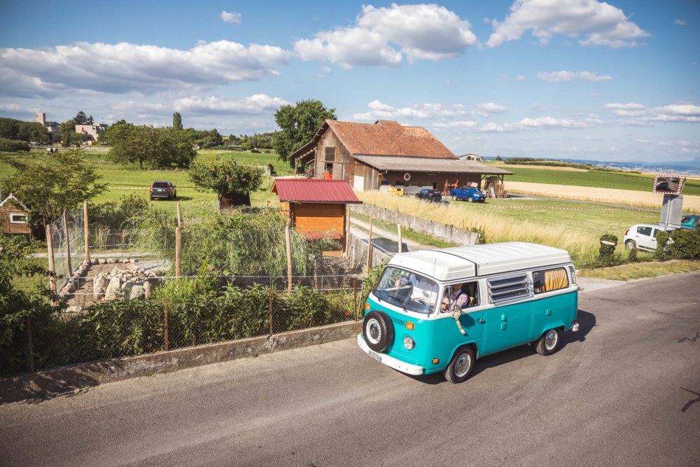 Une vieille camionnette VW bleue et blanche roule sur une route rurale à côté de champs et d'une maison au toit rouge et rouillé sous un ciel clair.