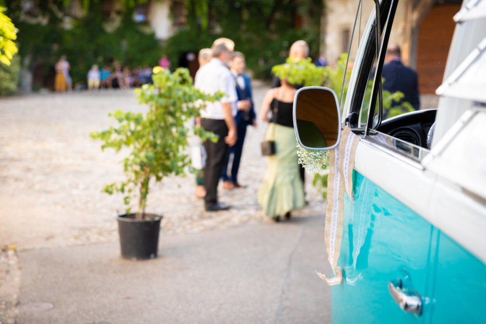 Une voiture bleue avec des rubans blancs est garée sur un lieu de mariage en plein air. Les invités se parlent en arrière-plan sur une surface de gravier.