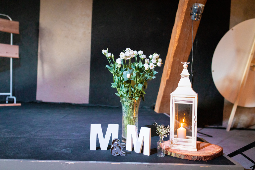 Une décoration de mariage de style champêtre se compose de fleurs blanches dans un vase, de grandes lettres « m m » blanches et d'une lanterne allumée sur une tranche de bois. Tous ces éléments sont placés sur une scène noire.