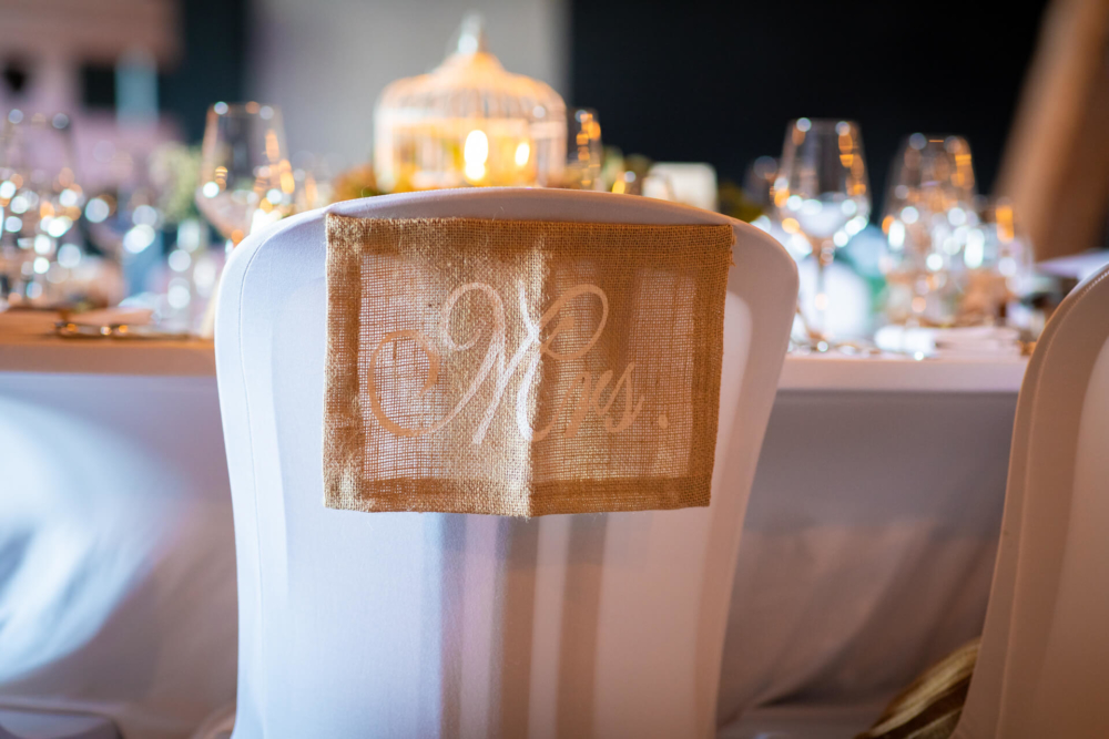 Une chaise de mariage fantaisie est décorée d'une housse blanche et d'un signe « mr » doré. La chaise est placée près d'une table décorée de porcelaine fine et de verrerie dans un lieu doucement éclairé.
