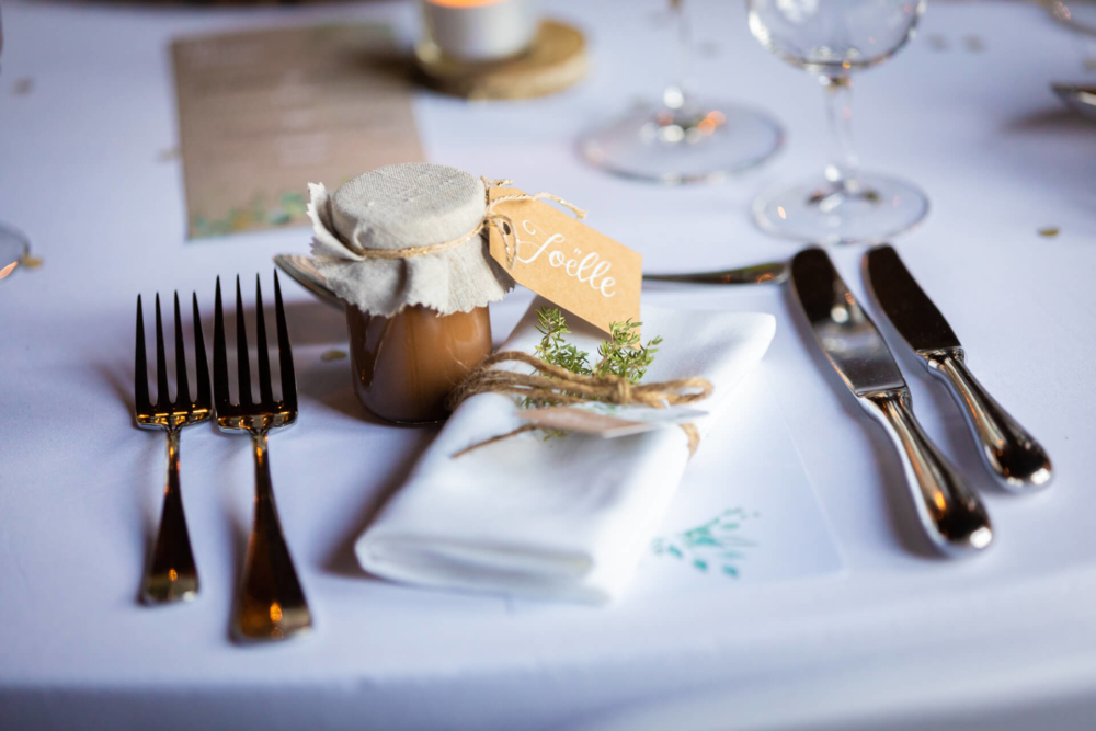 Un petit pot marqué « sicelle », une serviette blanche et des ustensiles en argent aux reflets dorés sont soigneusement disposés sur une nappe blanche pour un mariage élégant.