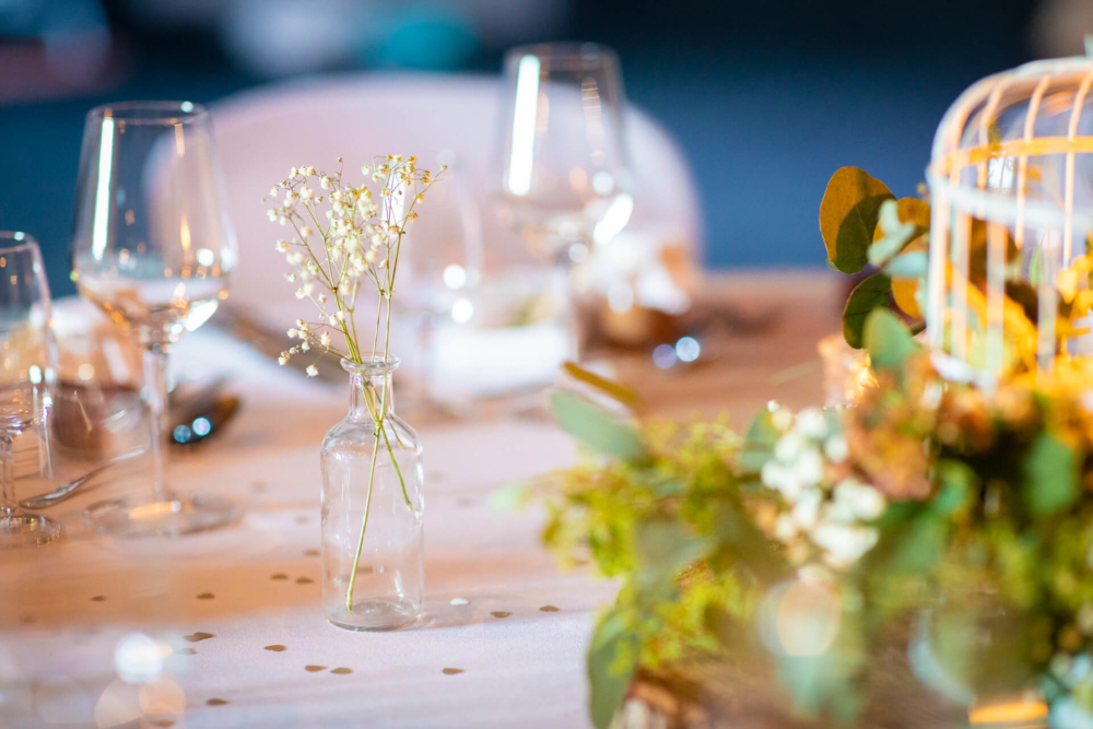 Une table à manger est joliment dressée avec des fleurs délicates dans un vase transparent, des verres en cristal et une lumière douce, créant une ambiance romantique.