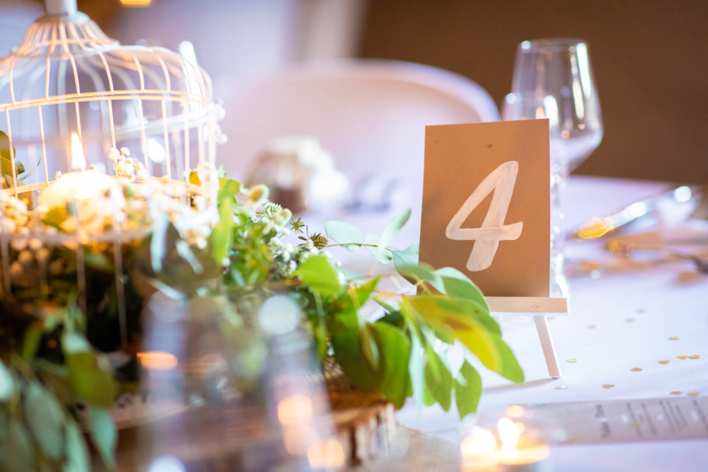 Une table de mariage numéro « 4 » est élégamment dressée avec une cage à oiseaux décorative entourée de fleurs blanches, des ustensiles de cuisine raffinés et un arrière-plan légèrement flou.