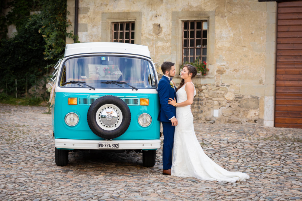 Un couple qui se marie s'embrasse devant une vieille camionnette Volkswagen turquoise et blanche garée dans une rue pavée à l'extérieur d'un bâtiment rustique.