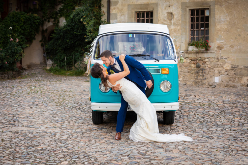Un couple récemment marié s'embrasse passionnément devant une vieille camionnette Volkswagen turquoise et blanche dans une rue pavée. La mariée porte une longue robe blanche et le marié est vêtu d'un costume bleu, tous deux se tiennent affectueusement.