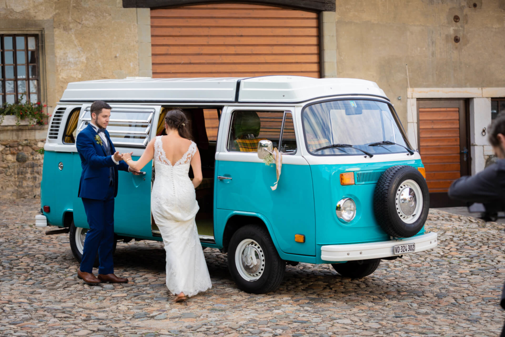 Un couple heureux se tient près d’une vieille camionnette Volkswagen turquoise et blanche, se tenant la main et se souriant. Un photographe prend leur photo.