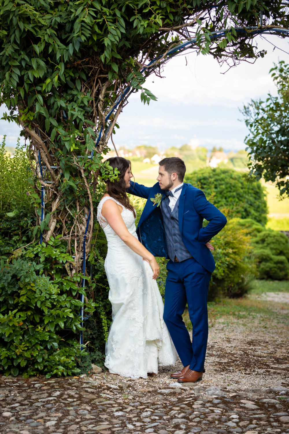 Une femme en robe de mariée blanche et un homme en costume bleu se tiennent sous une arcade avec des vignes, se souriant sur un chemin en pierre. Il y a un paysage vert derrière eux.