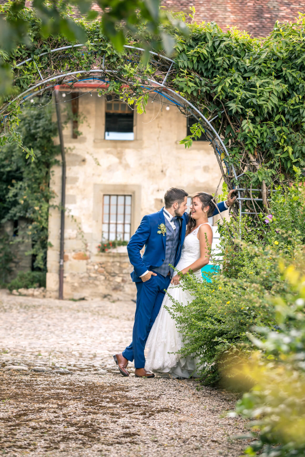 Des mariés souriants se trouvent sous une arche fleurie. Ils sont élégamment habillés, au milieu de plantes vertes, avec en arrière-plan une vieille bâtisse en pierre.