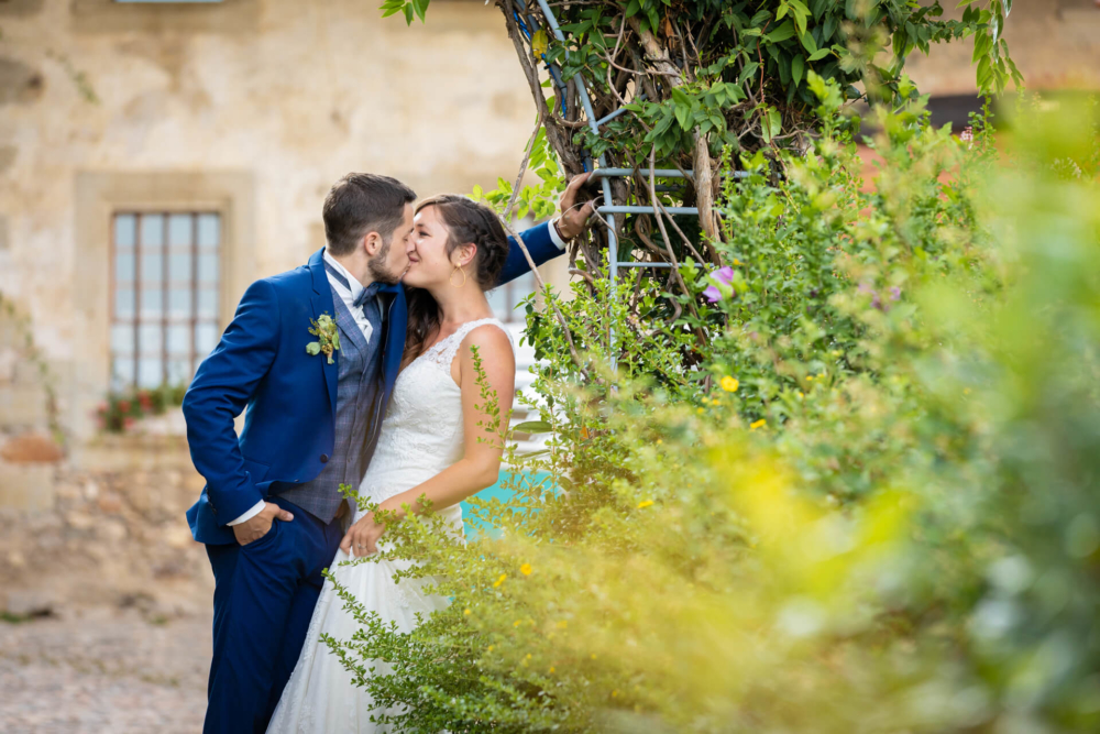 Un couple qui se marie s'embrasse intensément à côté d'une arche fleurie. Il y a des plantes vert vif devant et un vieux bâtiment en pierre derrière.