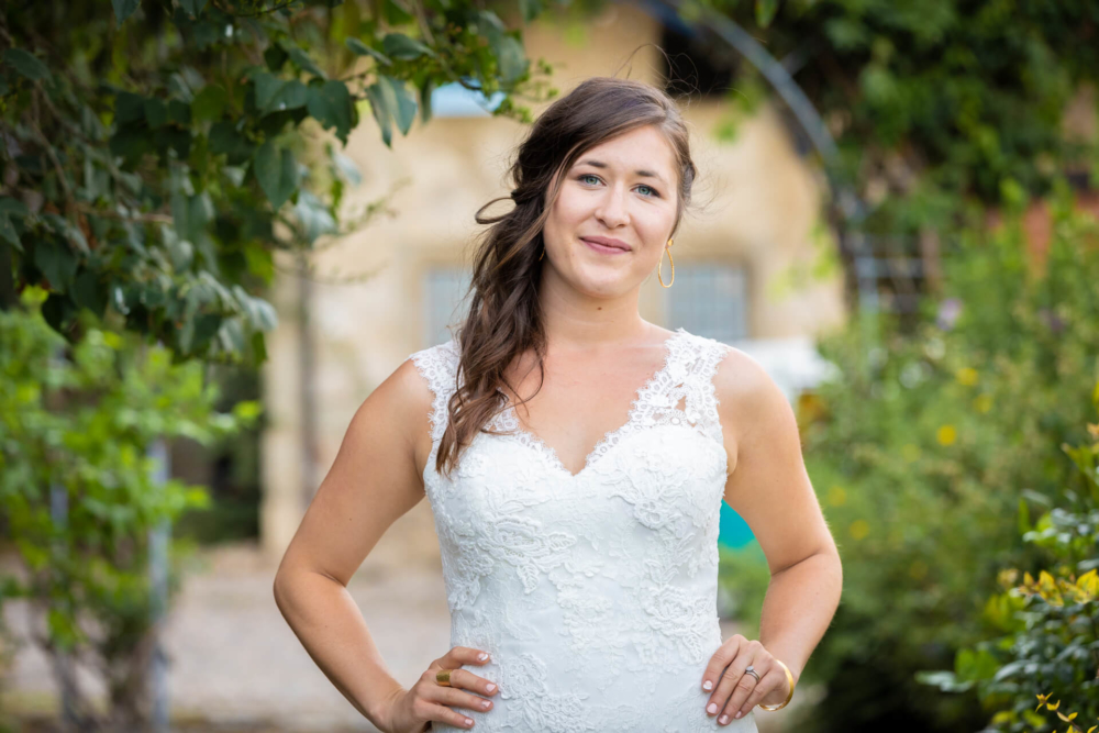 Une mariée souriante vêtue d'une robe de mariée en dentelle se tient dans un jardin. L'arrière-plan présente une verdure floue et un bâtiment.