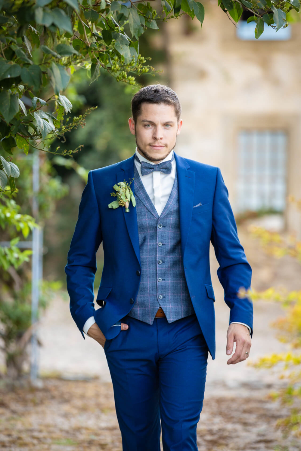 Un homme vêtu d'un costume bleu fantaisie, d'un gilet à carreaux et d'un nœud papillon se tient sous un arbre, regardant pensivement la caméra sur un arrière-plan flou.