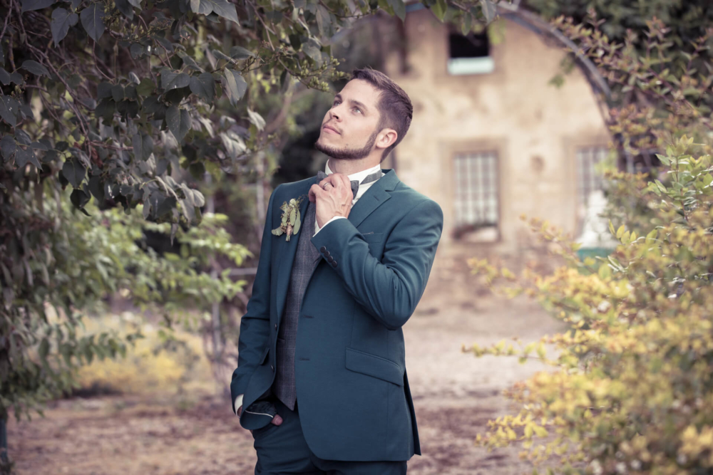 Un jeune homme réfléchi en costume vert attache sa cravate sous un arbre, avec un vieux bâtiment rural derrière lui.