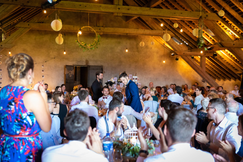 Une fête de mariage animée dans une grange aux poutres en bois est remplie d'invités élégamment habillés assis à des tables. Tout le monde applaudit tandis que le couple traverse la pièce.