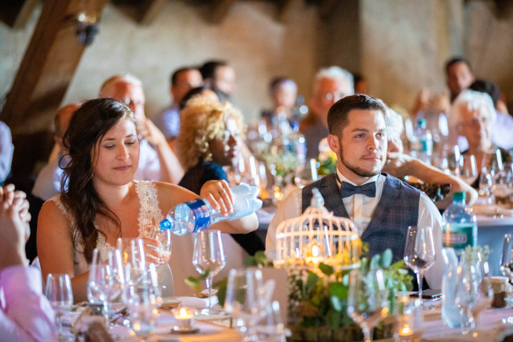 Un homme et une femme sont assis à une table de réception de mariage. La femme verse de l'eau et l'homme la regarde. Ils sont tous deux en tenue formelle, entourés d'un environnement festif.