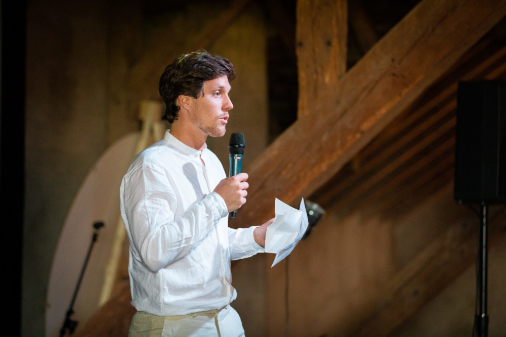 Un homme vêtu d’une chemise blanche tient des papiers et parle dans un microphone. Il se tient dans une salle en bois avec un éclairage agréable.