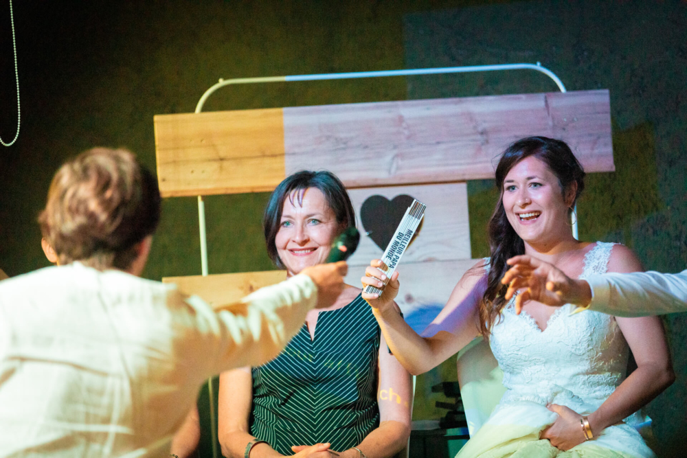 Une mariée et une autre femme profitent d'un moment de bonheur en riant avec un homme qui leur montre un billet surdimensionné dans un endroit très lumineux.