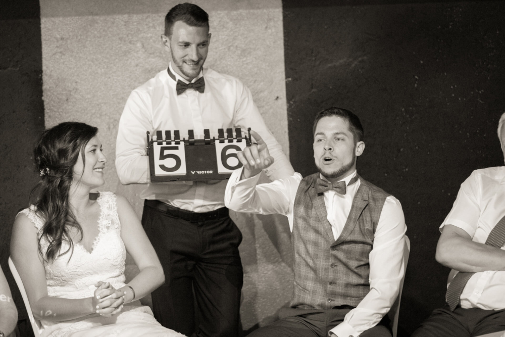 Les mariés sont vus sur une photo en noir et blanc avec deux témoins. L'un d'eux tient une ardoise de film intitulée « scène 5, prise 6 » pendant qu'il raconte une histoire. Ils sont tous habillés formellement et sourient.