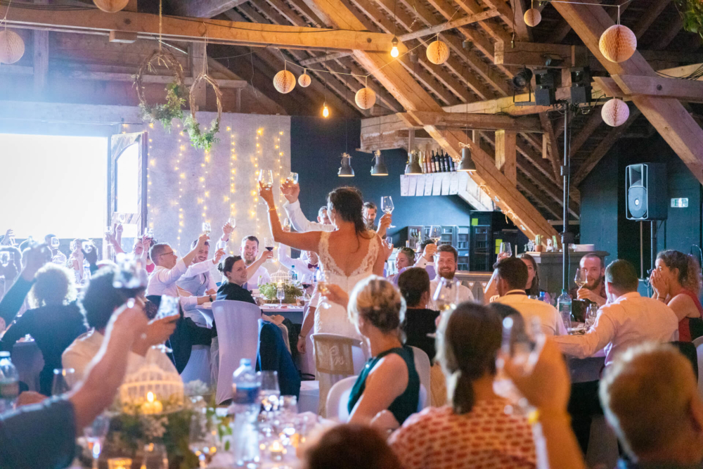 Un homme et une femme qui se marient lèvent leurs verres pour porter un toast dans une grange de style champêtre. La grange est décorée de guirlandes lumineuses scintillantes et de lanternes en papier. Leurs invités sont autour d’eux, les applaudissant et les acclamant.