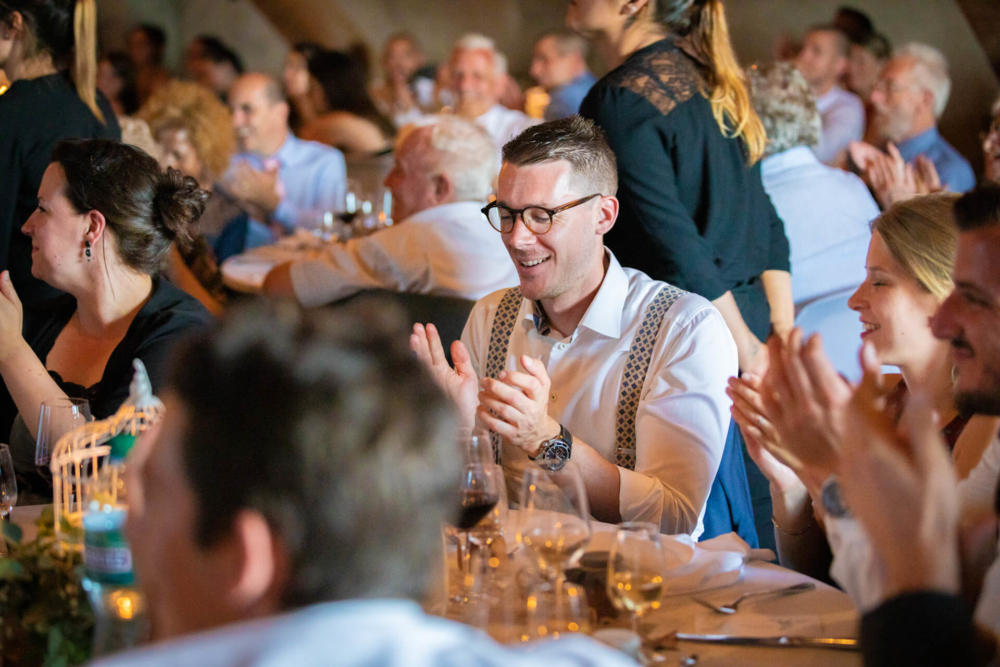 Un homme heureux vêtu de vêtements traditionnels et de lunettes applaudit lors d'un événement chargé avec de nombreux invités autour de lui, tous applaudissant également.