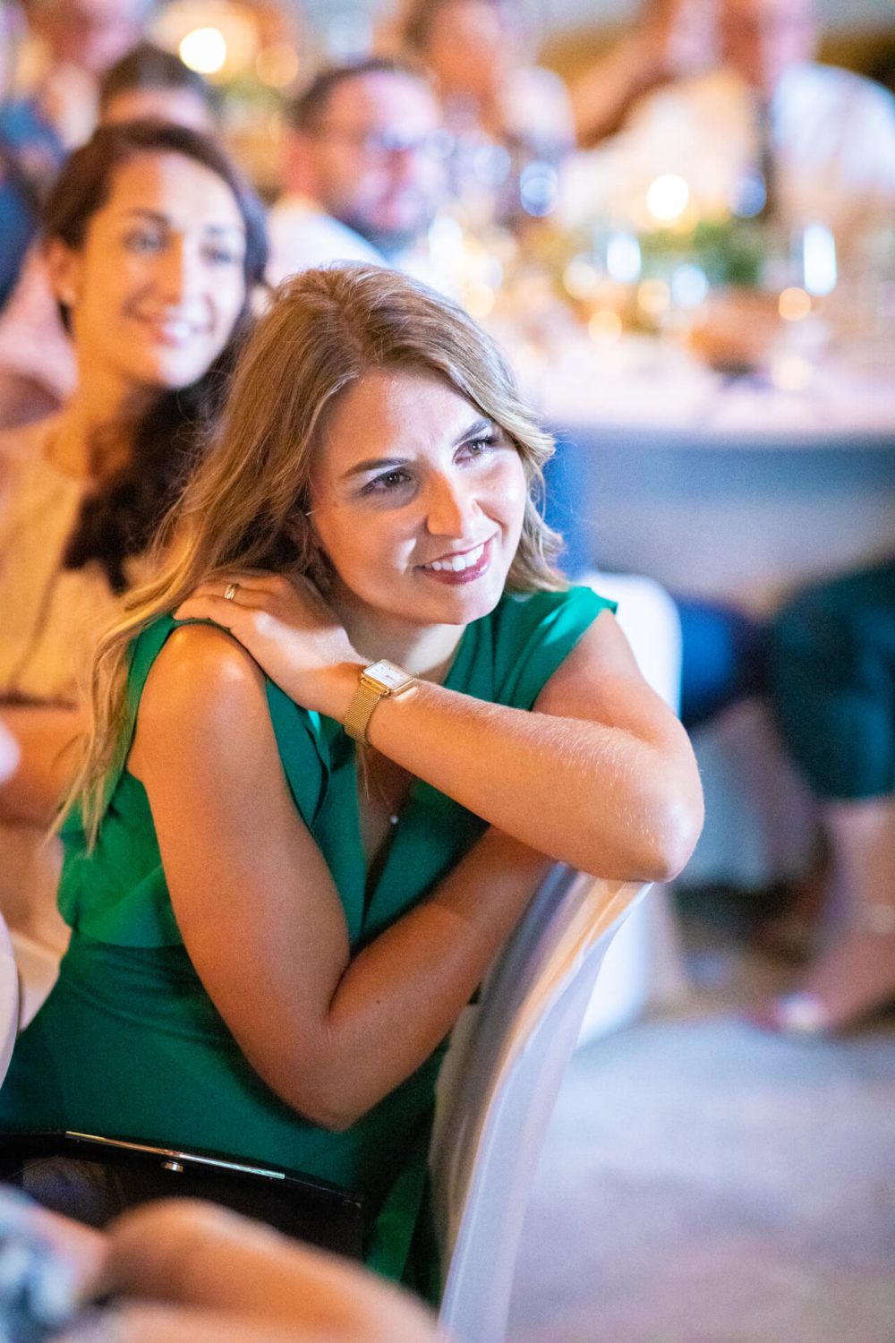 Une femme souriante vêtue d'une robe verte est assise avec d'autres invités lors d'un événement, regardant attentivement quelque chose que nous ne pouvons pas voir.