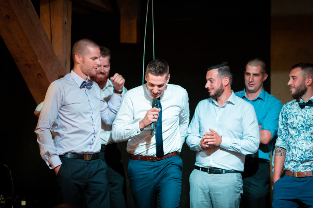 Un groupe d'hommes, habillés de façon semi-formelle, s'amusent autour d'un micro. L’un d’eux parle dans le micro tandis que les autres écoutent et sourient dans une pièce à la lumière chaleureuse.
