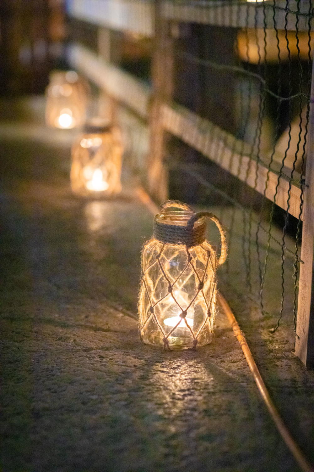 Une rangée de bougies dans des bocaux en verre enveloppés de ficelle est placée à côté d'un grillage, éclairant un chemin à peine visible au crépuscule.