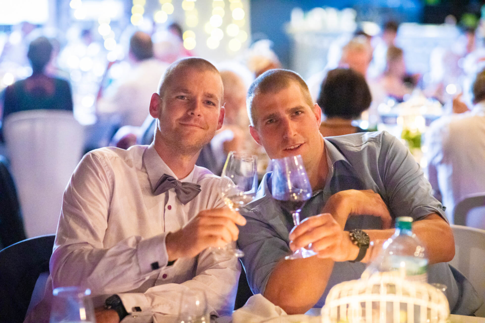 Deux gars habillés de manière semi-formelle, dont l'un porte un nœud papillon, brandissent joyeusement leurs verres à vin à une table de fête lors d'un événement nocturne.