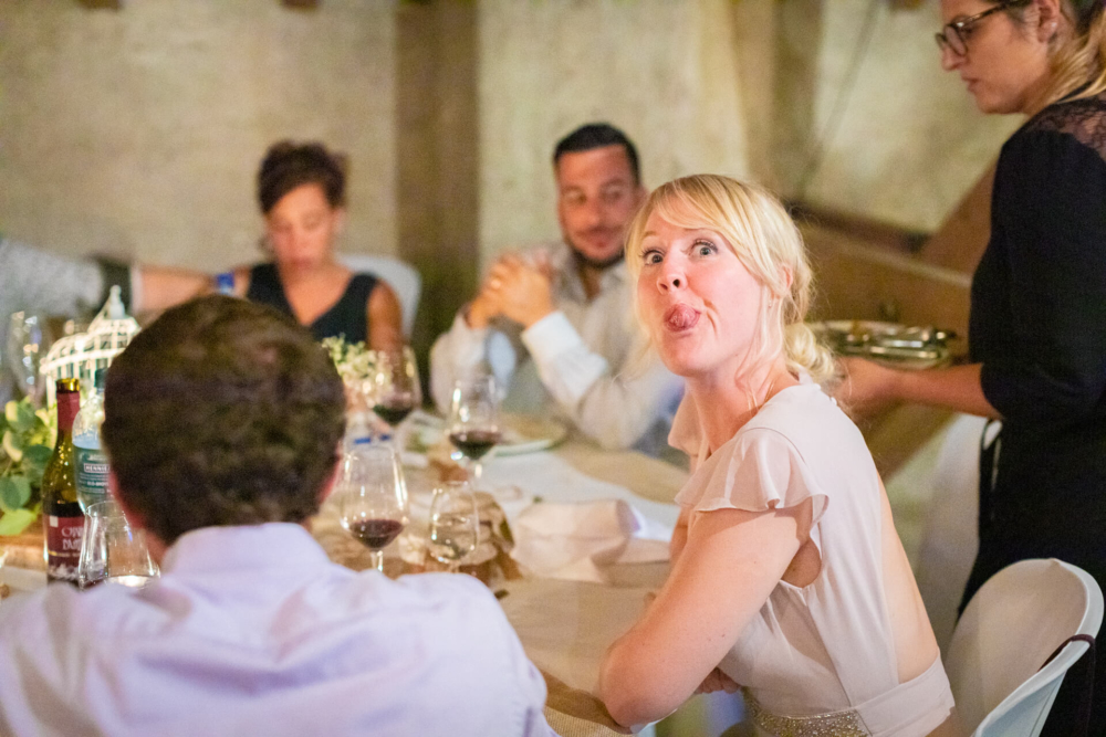Une femme vêtue d'une robe blanche tire la langue de manière ludique lors d'un dîner, pendant que les invités autour d'elle discutent.