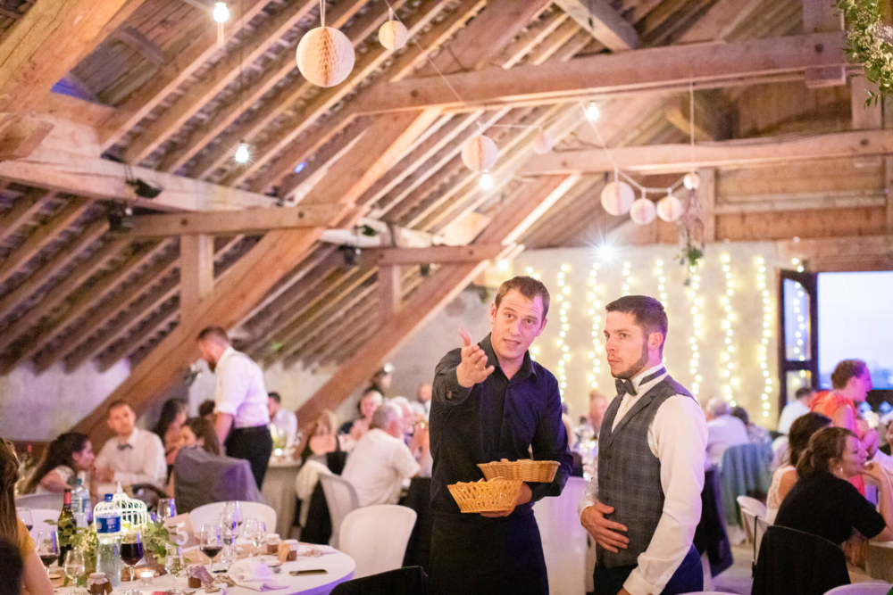 Deux hommes assistent à une simple réception de mariage dans une grange. Un homme tient un panier et parle avec des gestes tandis que l’autre écoute attentivement. Ils sont autour des invités et des guirlandes lumineuses.