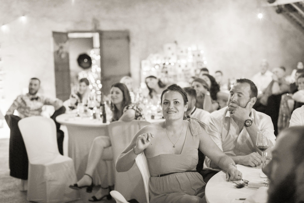 Un homme et une femme sont assis à une table lors d’une fête et prêtent attention à quelque chose devant eux. D'autres invités les entourent dans un endroit bien éclairé.