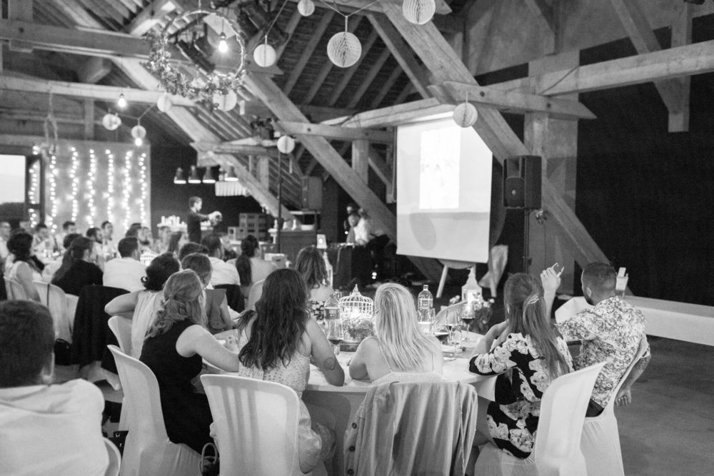 Les gens sont assis à des tables dans une grange de style champêtre et regardent une présentation sur un écran de projection. La grange a des guirlandes lumineuses et des poutres en bois au plafond.