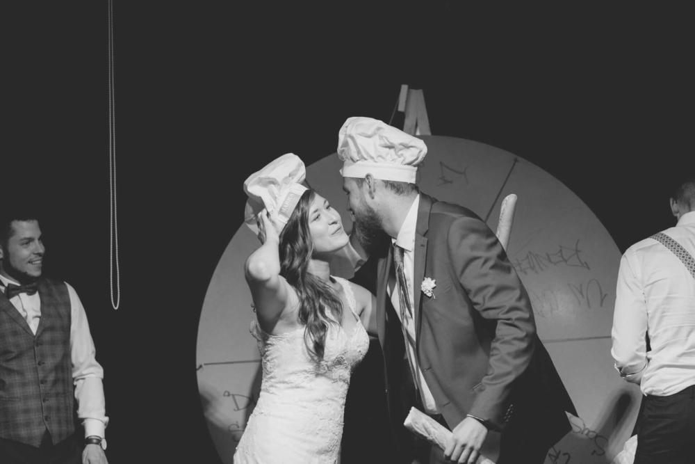 Une photo en noir et blanc montre un moment heureux lors d’une fête de mariage. Les mariés portent des toques de chef et se regardent avec amour, tandis que les invités à l'arrière sourient et s'amusent.