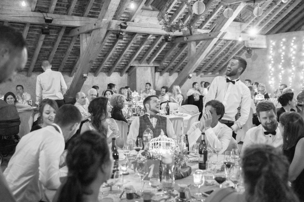 Une fête de mariage joyeuse et dynamique se déroule dans un lieu rustique avec des poutres en bois au-dessus. Les invités discutent et rient autour des tables décorées de plats élégants. Un homme en chemise blanche est debout, souriant largement.