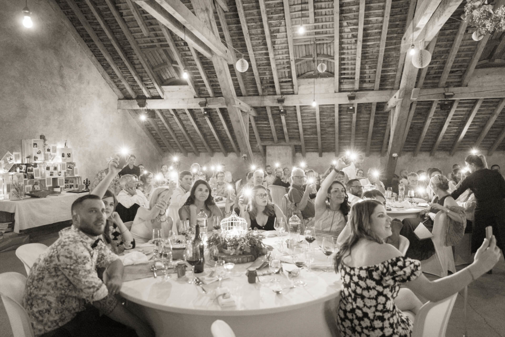 Une fête de mariage amusante dans une grange de campagne où les invités sont assis à des tables rondes, applaudissant et agitant les mains, éclairés par la douce lumière des ampoules suspendues.