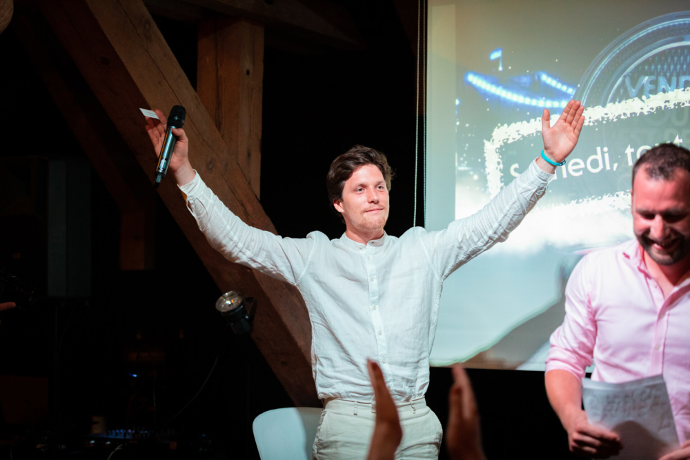 Un jeune homme heureux en chemise blanche est sur scène, levant les bras et tenant un microphone. Il y a un homme souriant à côté de lui et un public qui applaudit devant eux.