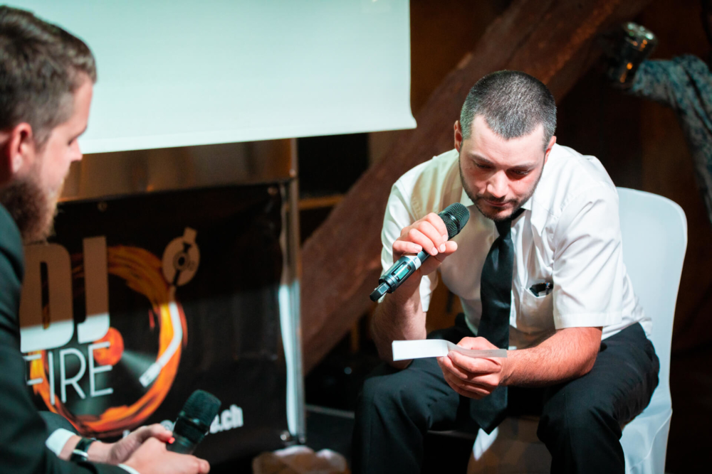 Un homme vêtu d’une chemise blanche et d’une cravate est assis sur un tabouret. Il tient un micro et une carte et parle sérieusement à un autre homme. L’autre homme dispose également d’un microphone et écoute attentivement. Ils sont à l'intérieur lors d'un événement.
