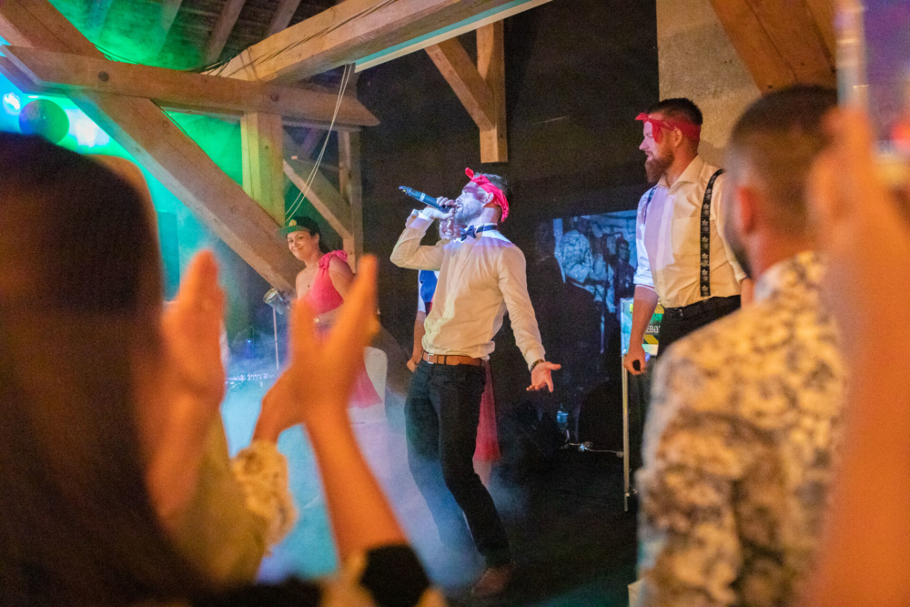 Un homme coiffé d’une casquette chante avec passion dans un microphone lors d’une fête animée aux lumières colorées. Le public applaudit et un membre du groupe regarde.