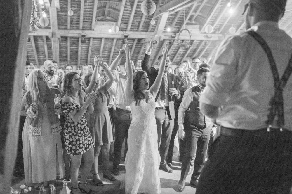 Une simple photo en noir et blanc montrant une joyeuse fête de mariage dans une grange. Les invités et la mariée lèvent joyeusement la main pendant qu'ils dansent. La photo est prise derrière quelqu'un qui regarde la foule.
