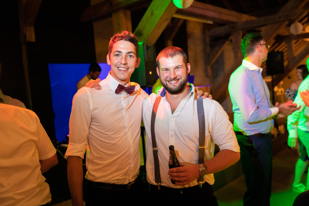 Deux hommes souriants lors d'une fête de mariage, l'un en chemise blanche et nœud papillon et l'autre en chemise blanche avec bretelles, tous deux tenant des bouteilles de bière.