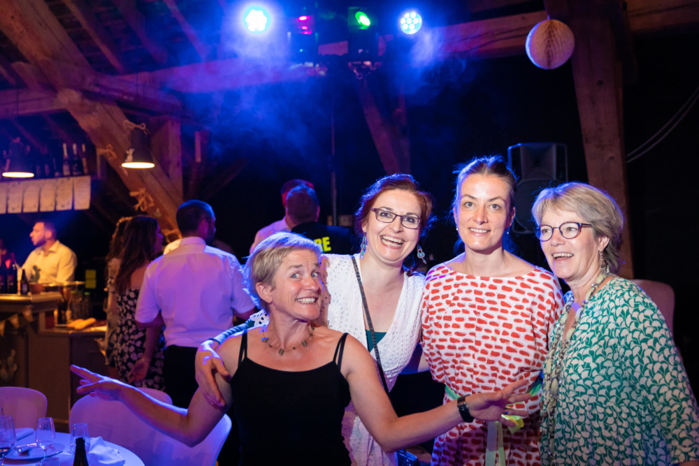 Quatre femmes participent joyeusement à une fête en salle avec des lumières vives. On aperçoit légèrement une foule derrière eux. Ils se tiennent proches et s’amusent clairement lors de la célébration.