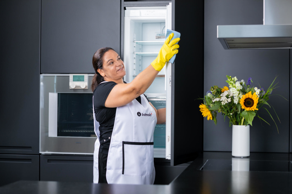Une femme vêtue d'un tablier blanc et de gants jaunes nettoie un réfrigérateur dans une cuisine moderne, avec des tournesols à proximité.
