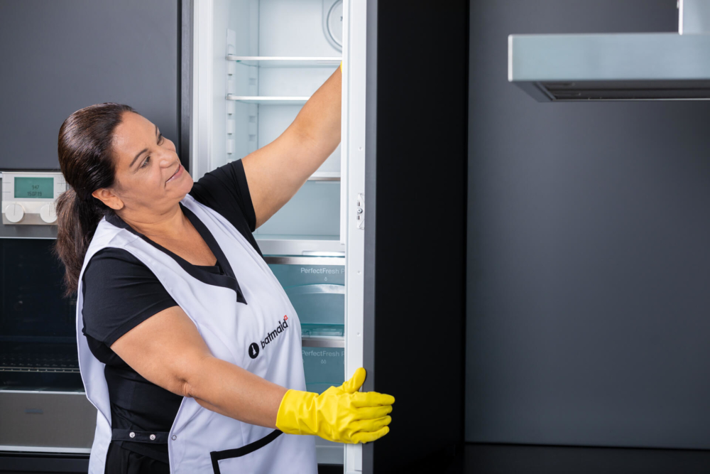 Une dame en uniforme et gants jaunes vérifie un réfrigérateur dans une cuisine contemporaine.