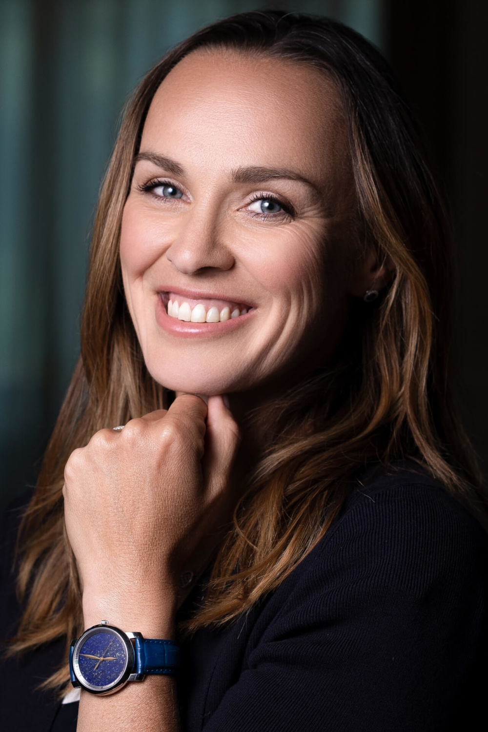 Martina-Hingis-Magazine-Regards-Beaurivage-Palace-Slika-ch-002 Une femme aux longs cheveux bruns sourit, vêtue d'un chemisier foncé et d'une montre bleue. Son menton repose sur sa main.