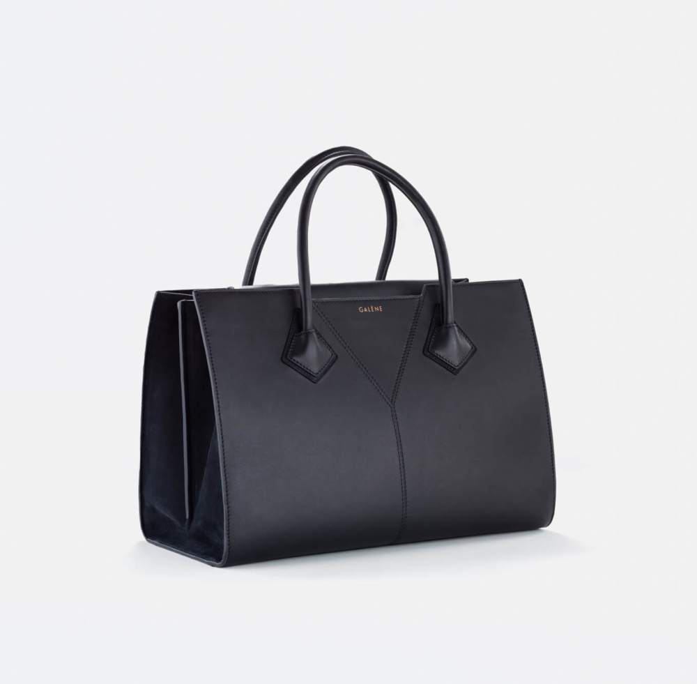 Un sac fourre-tout simple en cuir noir avec des poignées droites et des coutures visibles. Le nom de la marque est gravé au milieu, sur un fond gris clair.