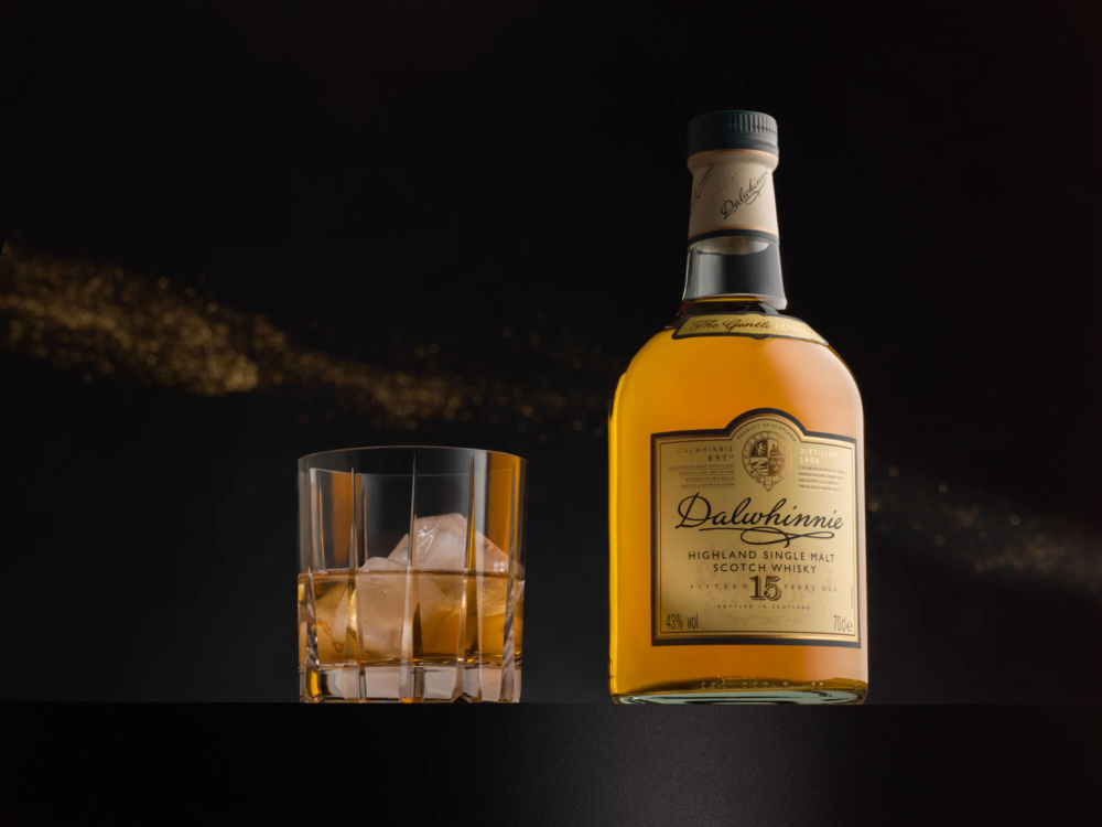 Une bouteille de whisky écossais single malt Dalwhinnie Highland se trouve à côté d'un verre avec des glaçons. Le tout sur un fond sombre avec de douces paillettes dorées.