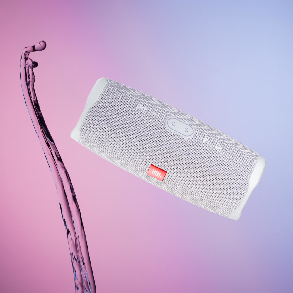 Un haut-parleur Bluetooth portable JBL blanc se trouve sur un fond dégradé violet et rose, avec un liquide violet foncé coulant à côté.