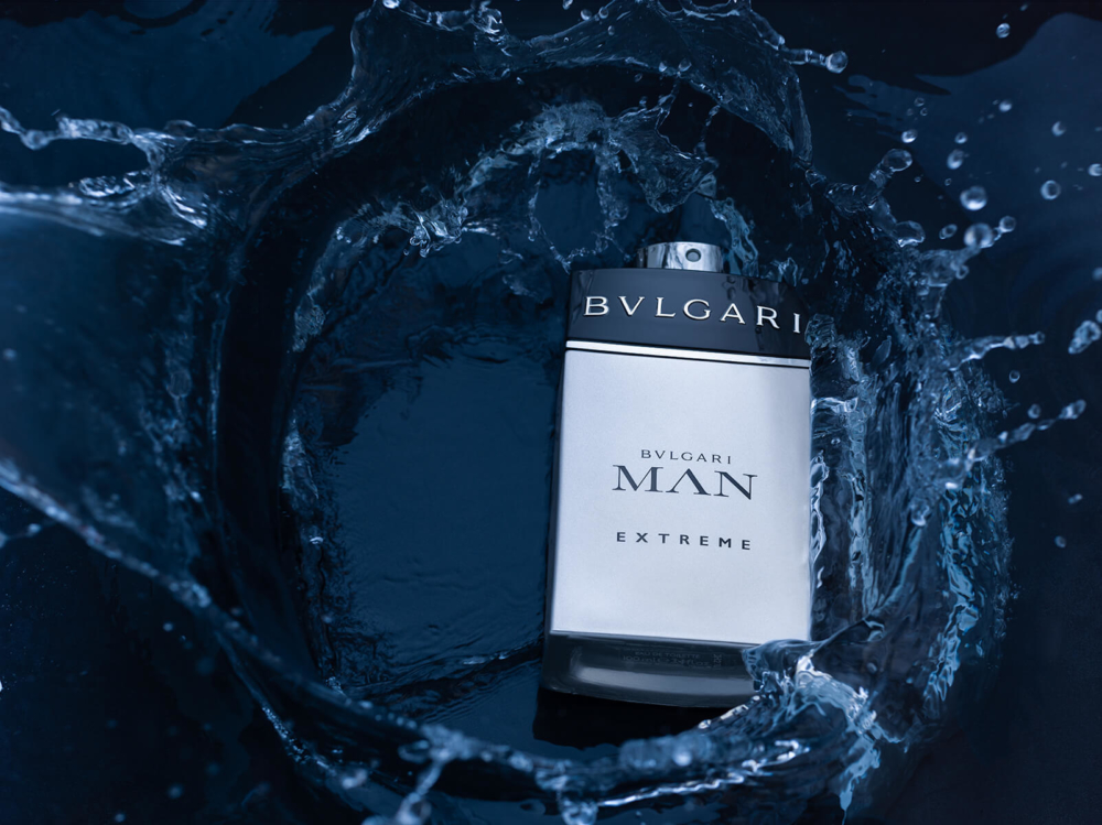 Une photo montre une bouteille d’eau de Cologne Bvlgari Man Extreme au milieu d’éclaboussures d’eau, sur un fond bleu foncé. L’image dégage une sensation vibrante et luxueuse.
