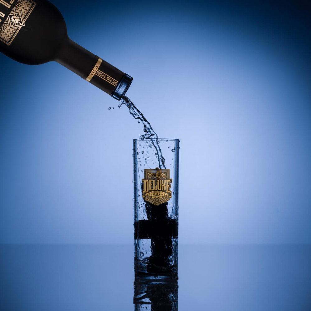 Une bouteille sombre étiquetée « de luxe » verse un liquide clair dans un verre assorti sur fond bleu, montrant l'éclaboussure vibrante.